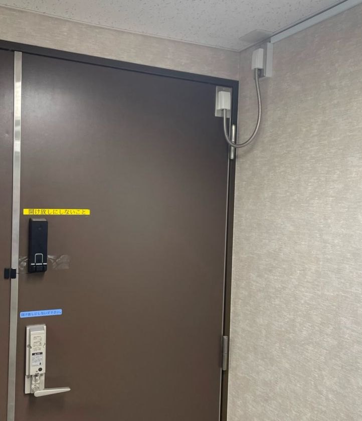 面付けの通電金具での施工例の写真（室内側）。扉の上部の枠と扉に通電金具、枠側の金具から天井へモールで配線、扉の中ほどに RemoteLOCK のインナーユニット